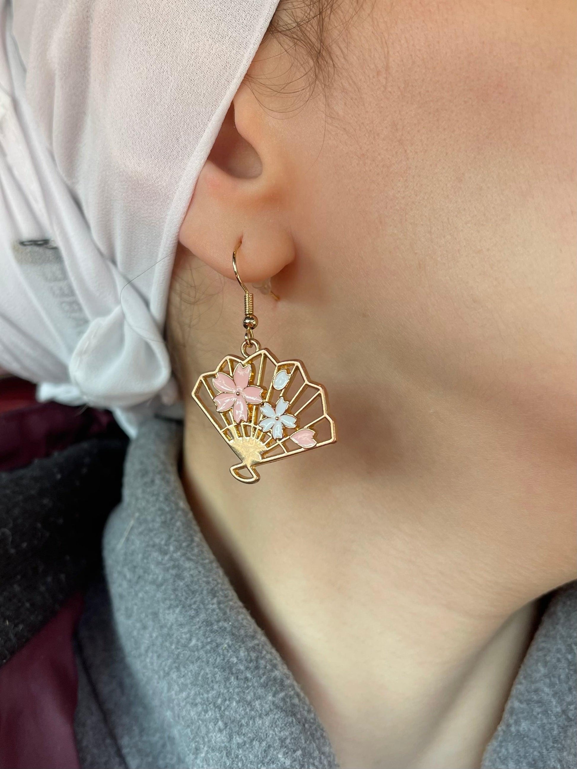 Close up on an ear with asymmetric asymmetrical dangle dangling drop fashion jewelry earrings, with a gold base traditional japanese fan charm and sakura flower - Roulette Safari